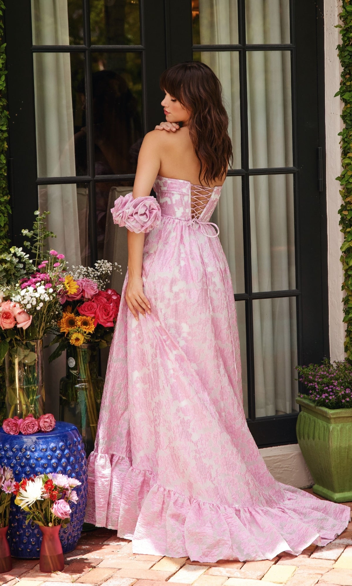 A woman wearing the Ava Presley 40035 long formal dress stands with her back to the camera, surrounded by potted flowers and plants near glass doors.